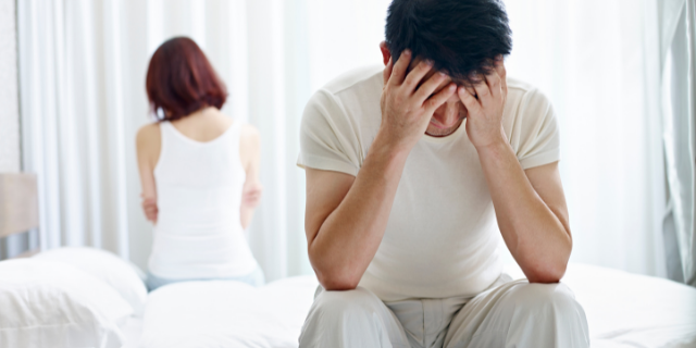 Homem e mulher sentados na beira da cama, um de costa para o outro. O homem está com as duas mãos na cabeça