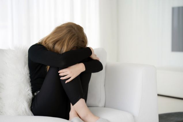 Imagem de uma mulher sentada e com a cabeça baixa em um sofá branco toda de preto