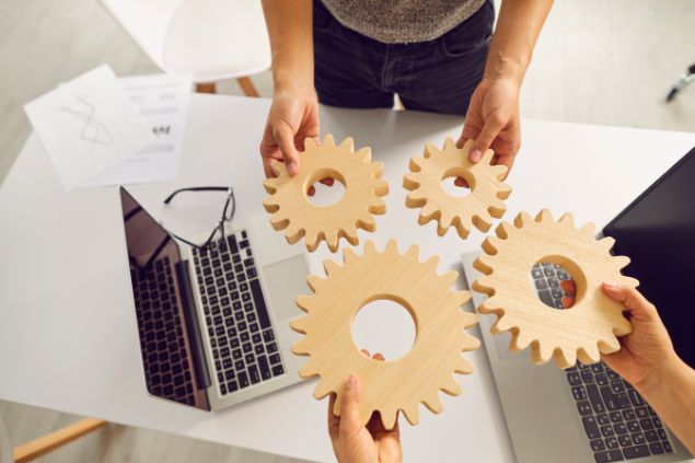 Imagem de duas pessoas em um escritório, cada um asegurando duas engrenagens feitas de MDF e laptops na mesa