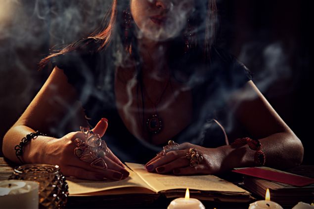 Mulher com um livro a sua frente, velas ao redor da mesa em que está sentada e em um ambiente meia-luz, como se estivesse fazendo a previsão de algo para alguém 