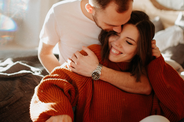 Homem abraçando sua parceira por trás e dando um beijo em sua teste enquanto ela sorri
