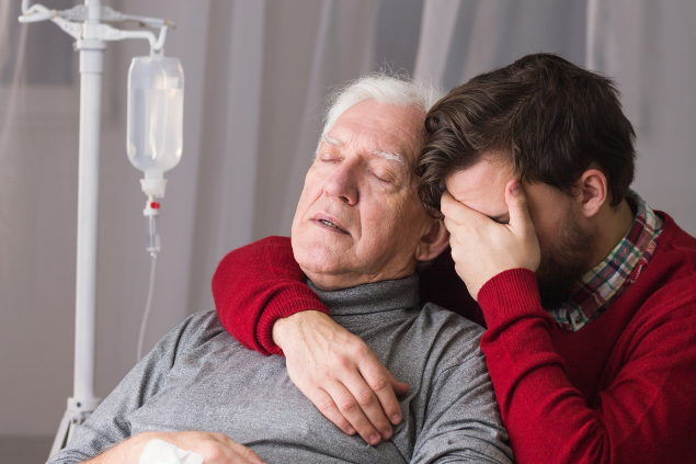 Homem mais velho deitado tomando soro enquanto filho está ao lado dele com as mãos no rosto