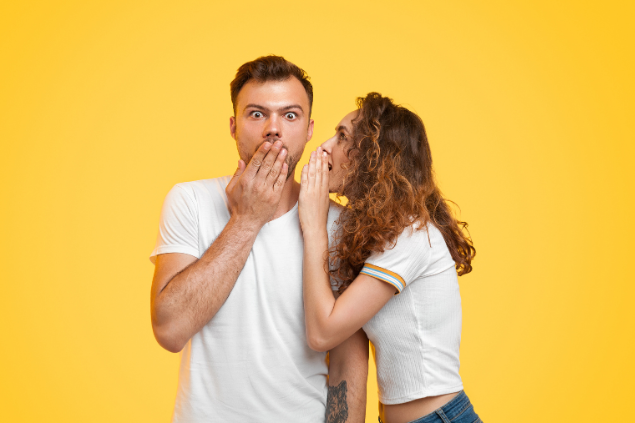 Uma mulher falando no ouvido de um homem, enquanto ele está com feição de surpreso 