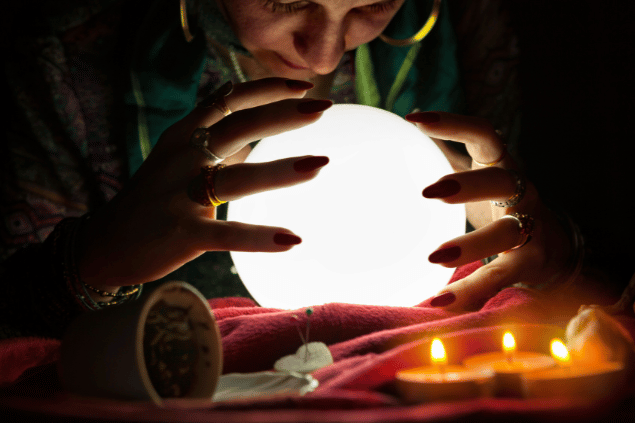 Uma cartomante está concentrada e com as duas mãos ao redor de uma bola de cristal.