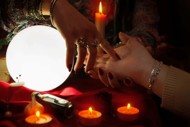 Uma cartomante faz a leitura da palma da mão de uma cliente. As mãos da cartomante e da consulente estão no foco da imagem.