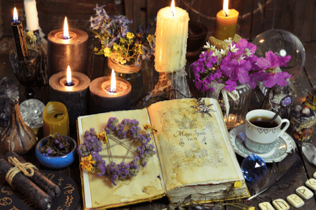 Existem vários objetos em uma mesa de madeira, como: um livro aberto com um pequeno arranjo de flores em cima, uma xícara com um líquido dentro, recipientes com flores, uma bola de cristal e muitas velas acesas ou apagadas.