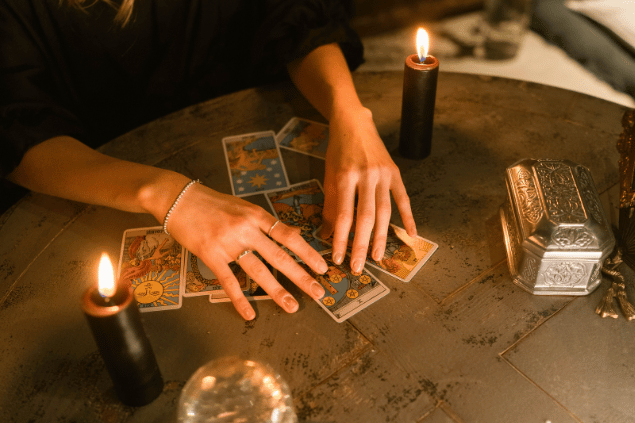 As mãos de uma mulher mexendo em algumas cartas de tarot estão no foco da imagem. Ao lado, há velas acesas na mesa.