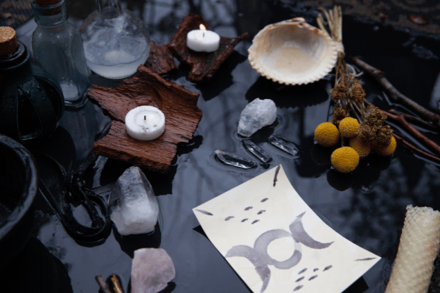 Há vários itens sobre uma mesa de madeira molhada pela chuva, como: cristais, uma planta, uma concha, velas, um papel e garrafas de vidro.