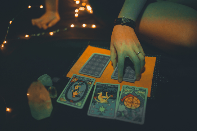 A mão de uma mulher está mexendo em um baralho de tarot que está sobre uma mesa. Ao lado, há cristais na mesa.