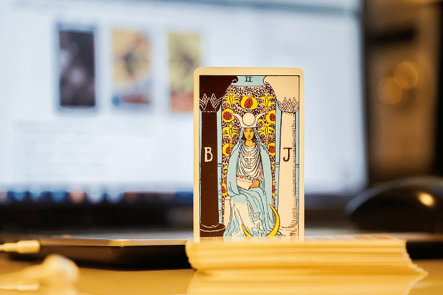 Na imagem, há a carta A Sacerdotisa do Tarot na posição normal e em pé. Ao fundo, há um notebook aberto.