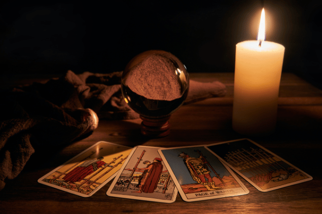 Quatro cartas de Tarot estão dispostas sobre uma mesa de madeira. Ao lado, há uma bola de cristal e uma vela.