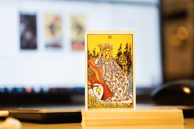 Na imagem, há a carta A Imperatriz do Tarot na posição normal e em pé. Ao fundo, há um notebook aberto.