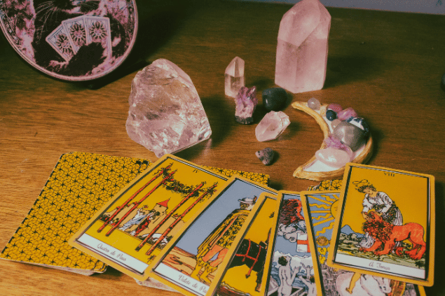 Algumas cartas de Tarot estão dispostas ao lado de um baralho sobre uma mesa. Há também cristais e um quadro.