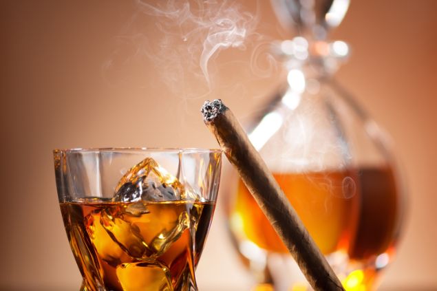Imagem de uma garrafa e copo com cachaça e ao lado, um cigarro aceso, simbolizando uma oferenda para o Exu Veludo.