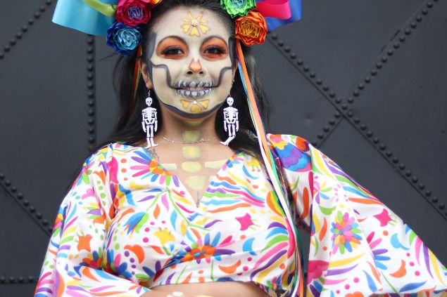 Imagem de uma mulher com uma pintura de caveira no rosto. Ela usa roupas coloridas e uma tiara feita de flores coloridas, simbolizando a pombagira Rosa Caveira.