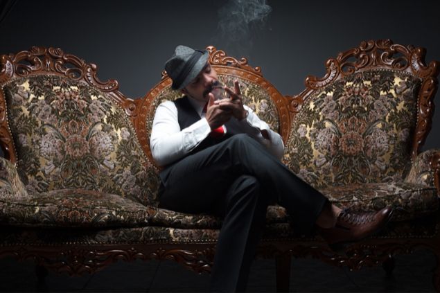 Imagem de um homem sentando em uma poltrona. Ele usa um chapeu estilo malandro e fuma um charuto, simbolizando a entidade Zé Malandro na Umbanda.