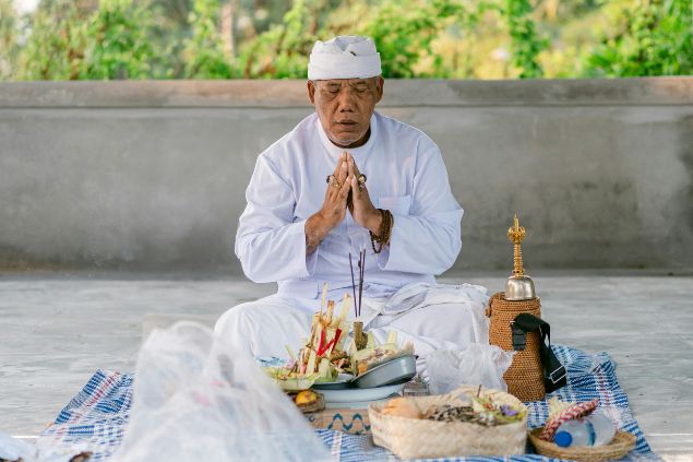Imagem de um homem usando uma roupa e um turbante na cor branca. Ele está sentando no chão, rezando e com algumas oferendas dispostas sobre um pano, simbolizando o o Babalorixá no candomblé.