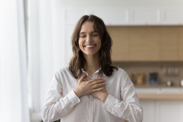 Imagem de uma mulher usando uma camisa branca, ela está sorrindo e feliz, com os olhos fechados, com as mãos sobre o peito, simbolizando o gesto de gratidão.