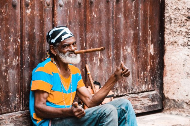 Imagem de um homem preto, sentado no chão fumando um cachimbo, simbolizando o pai Maneco da Umbanda.