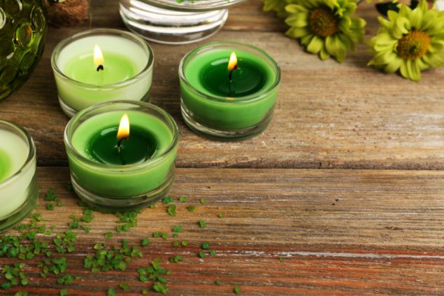 Imagem de várias velas acesas na cor verde, dispostas sobre uma mesa de madeira, decorada com algumas folhas verdes, simbolizando uma das oferendas para Zé Pereira.