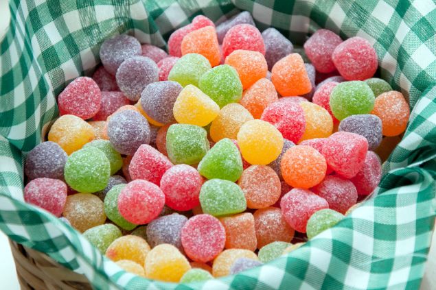 Imagem de uma cesta com tecido xadrez nas cores verde e branco, contendo balas de jujuba coloridas, uma das oferendas para o Erê Joãozinho da Umbanda.