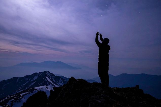 Imagem da silhueta de um homem em cima de uma montanha com as mãos elevadas, em sinal de oração.