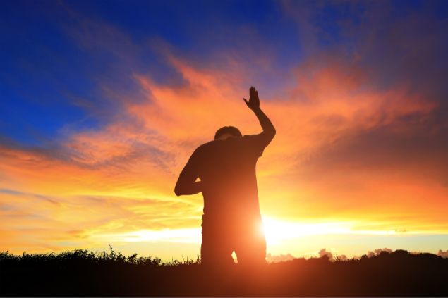 Imagem de fundo de um lindo pôr do Sol e em destaque um homem ajoelhado com uma de suas mãos para o alto, em oração.