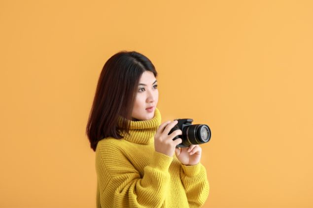 Imagem de fundo amarelo e em destaque uma mulher fotógrafa segurando uma câmera em mãos.