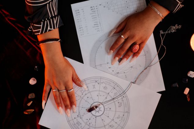 Imagem de duas folhas com os mapas astrais desenhados nelas. Sobre eles, as mãos de uma astróloga.