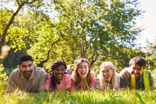Imagem de um campo verde e em destaque vários jovens deitados sobre o gramado e todos estão sorrindo.