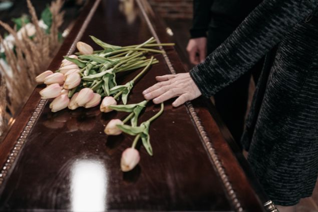 Imagem de uma mão sobre um caixão. Nele estão dispostas algumas flores de tulipa branca. A imagem simboliza o funeral de uma pessoa desconhecida.