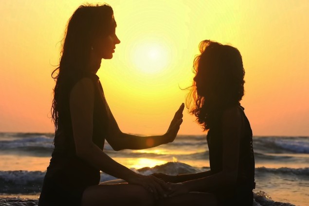 Imagem de um lindo pôr do Sol em uma praia e em destaque duas mulheres sentadas na água do mar, uma de frente para a outra, sendo que uma delas está emanando energia para a outra com uma de suas mãos.