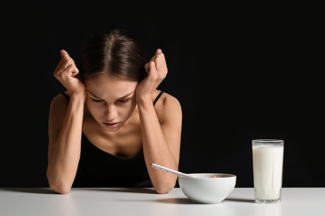 Imagem de uma mulher com as mãos sobre a cabeça. Ao lado dela, um copo com leite e uma tigela com cereais. Ela sofre com problema de anorexia.