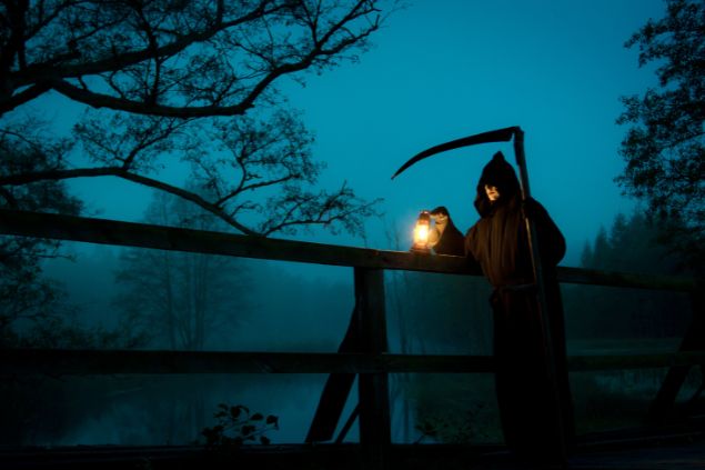 Imagem de uma noite nebulosa e em destaque um homem com a roupa da morte segurando em uma das mãos uma foice e na outra uma lanterna. Ele está parado em uma ponte velha