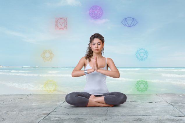 Imagem de uma jovem fazendo meditação na praia. Em volta dela, os elementos de cada um dos 7 chakras brilhando ao seu redor