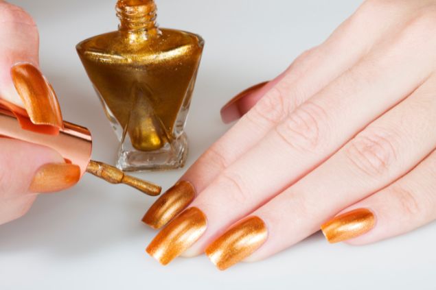 Imagem de uma mão feminina com as unhas sendo pintadas com esmalte na cor dourada.