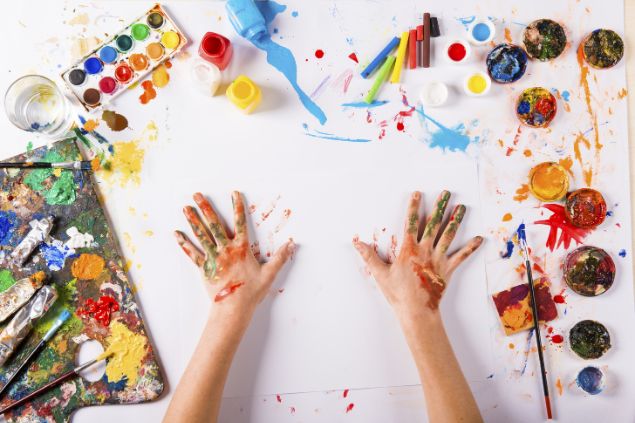 Imagem das mãos de uma pessoa sobre uma mesa com papel branco, tintas e pincéis para fazer uma aula de artes plásticas, uma das habilidades do signo de peixes.