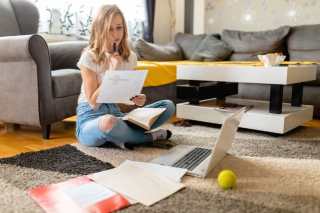 Imagem de uma mulher do signo de sagitário sentada no chão da sala, com uma carinha que não gosta muito de estudar. Ao lado do computador uma bolinha de tênis.