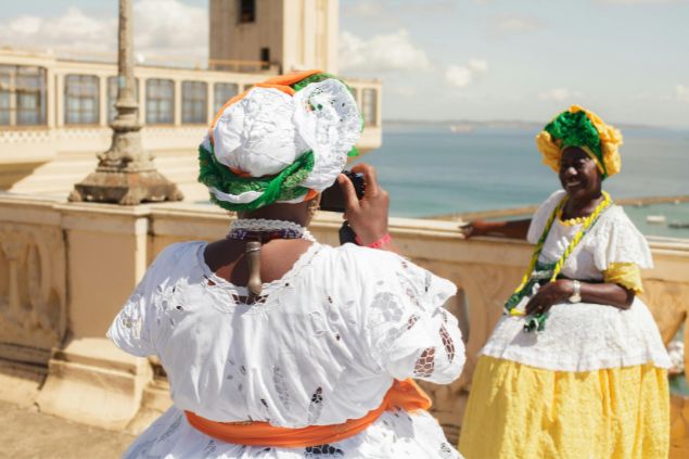 Imagem de duas baianas no elevador de Salvador. Uma delas está pousando para foto e ao fundo dela, o mar.