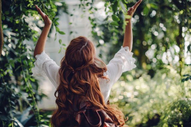 Imagem de uma mulher de cabelos longos avermelhados em um jardim com mas mãos erguidas. A foto traz o conceito de conexão vibratória com as entidades espirituais da Umbanda.