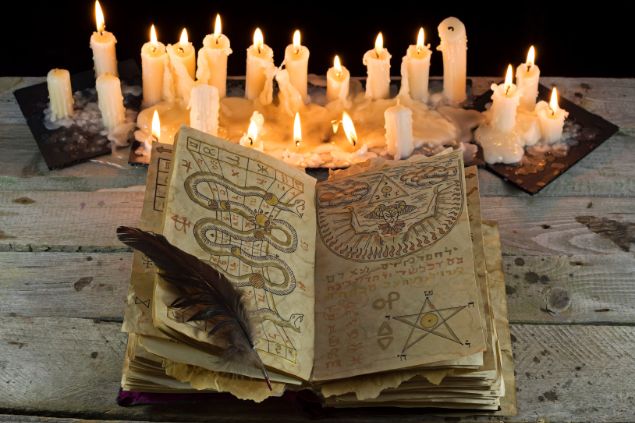 Imagem de um livro com diversos tipos de pontos riscados. Ele está aberto e sobre ele, uma pena. Ao fundo, várias velas brancas acesas.