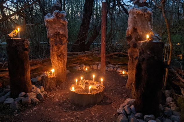 Imagem de um despacho feito com muitas velas acesas em um jardim. O cenário conta ainda com troncos de árvores e uma ossada de animal.