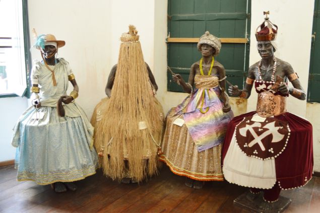 Imagem de vários tipos de orixás feitos de cera, expostos em uma sala.