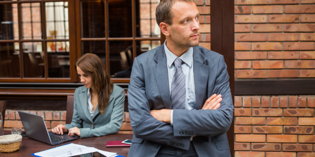 Homem de braços cruzados com semblante. Ao fundo, há uma mulher sentada.