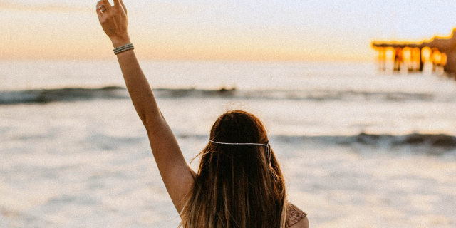 Mulher com o braço erguido anda em direção ao mar.