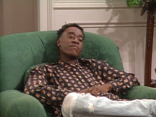 Homem negro sentado numa poltrona verde, usando camisa estampada e calça rasgada.