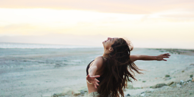 Mulher sorrindo com as costas curvadas e braços erguidos. Ela está em cenário externo.