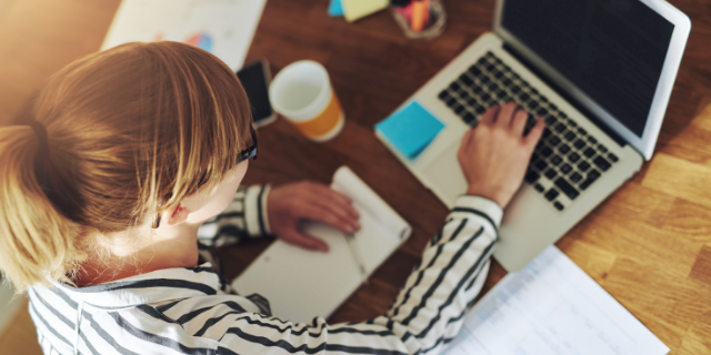 Mulher digita em computador sobre uma mesa. Há também papéis, um celular e um copo de plástico. 