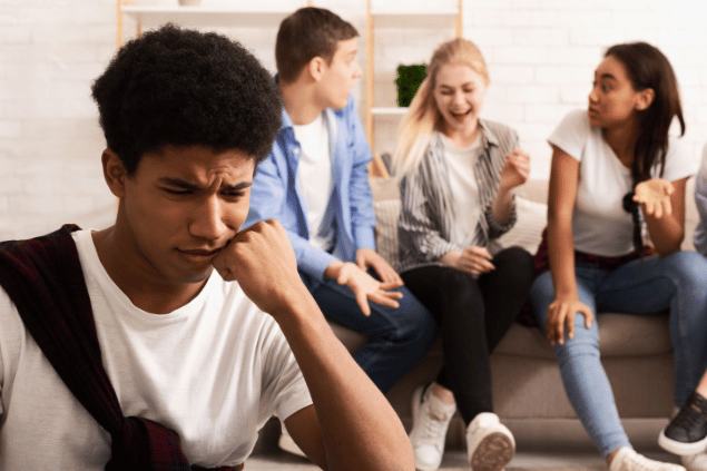 Menino sentindo-se excluído pelo seu grupo de colegas, enquanto eles conversam ao fundo. 