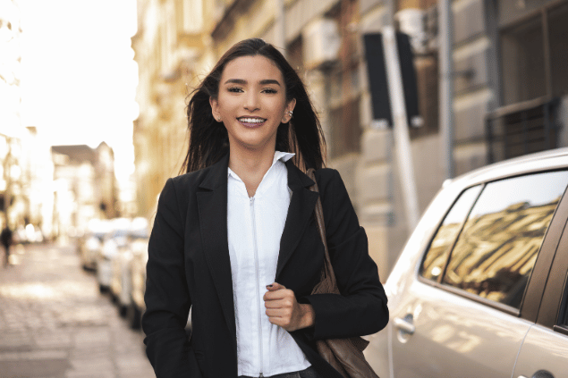 Pollyana Ventura / Getty Images Signature / Canva Uma mulher de cabelos castanhos que veste roupas formais está sorrindo. A imagem sinaliza que a mulher está no mercado de trabalho e é independente. Ao fundo, há uma cidade desfocada.